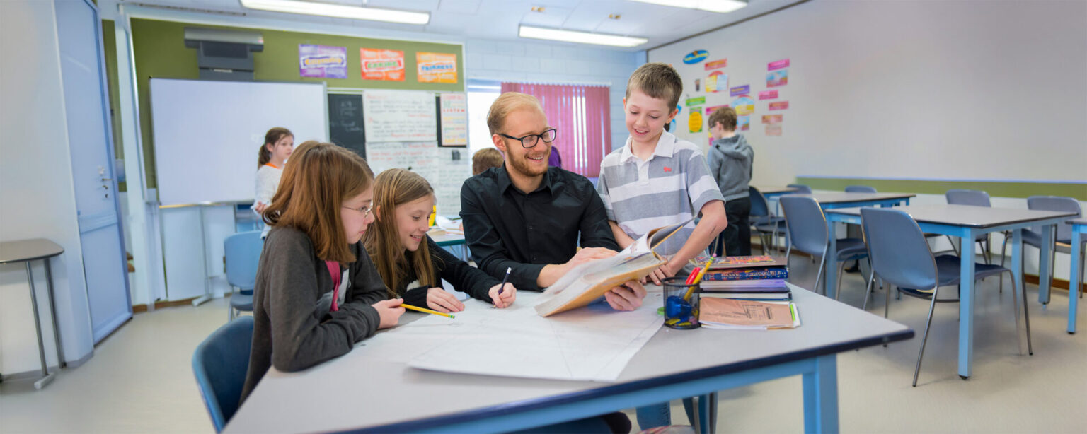 A teacher in black shirt reading a book with a student while 2 other girls sit with them in a class room setting