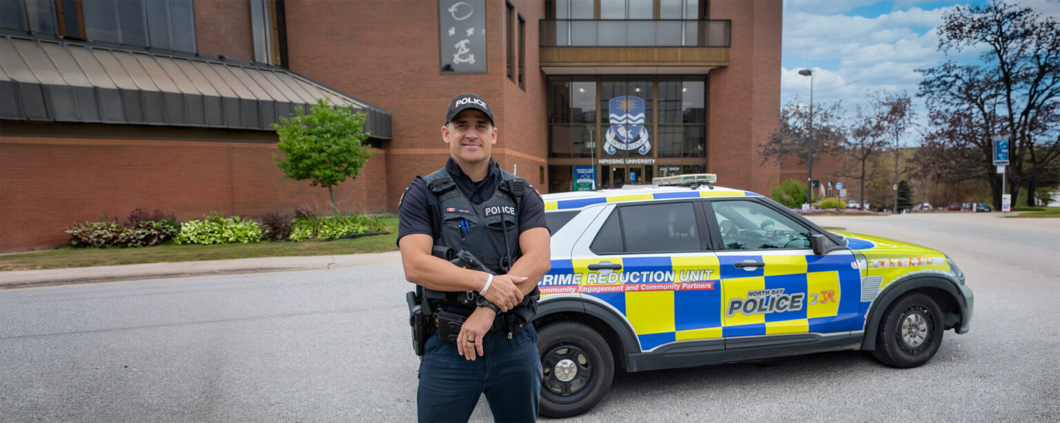 A police officer next to his police car in front of the Nipissing campus entrance
