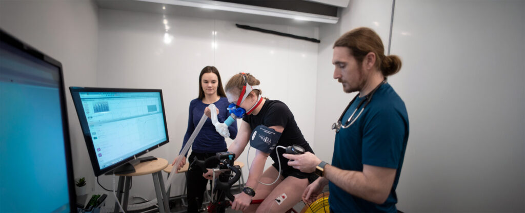 2 students running some test on another student on a bike connected to wires in a lab