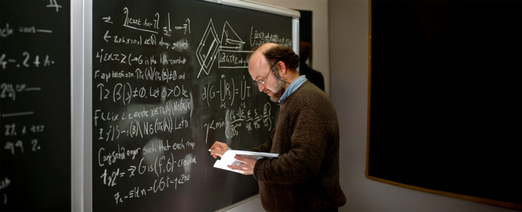 A professor in brown sweater writing on a black board with white equations