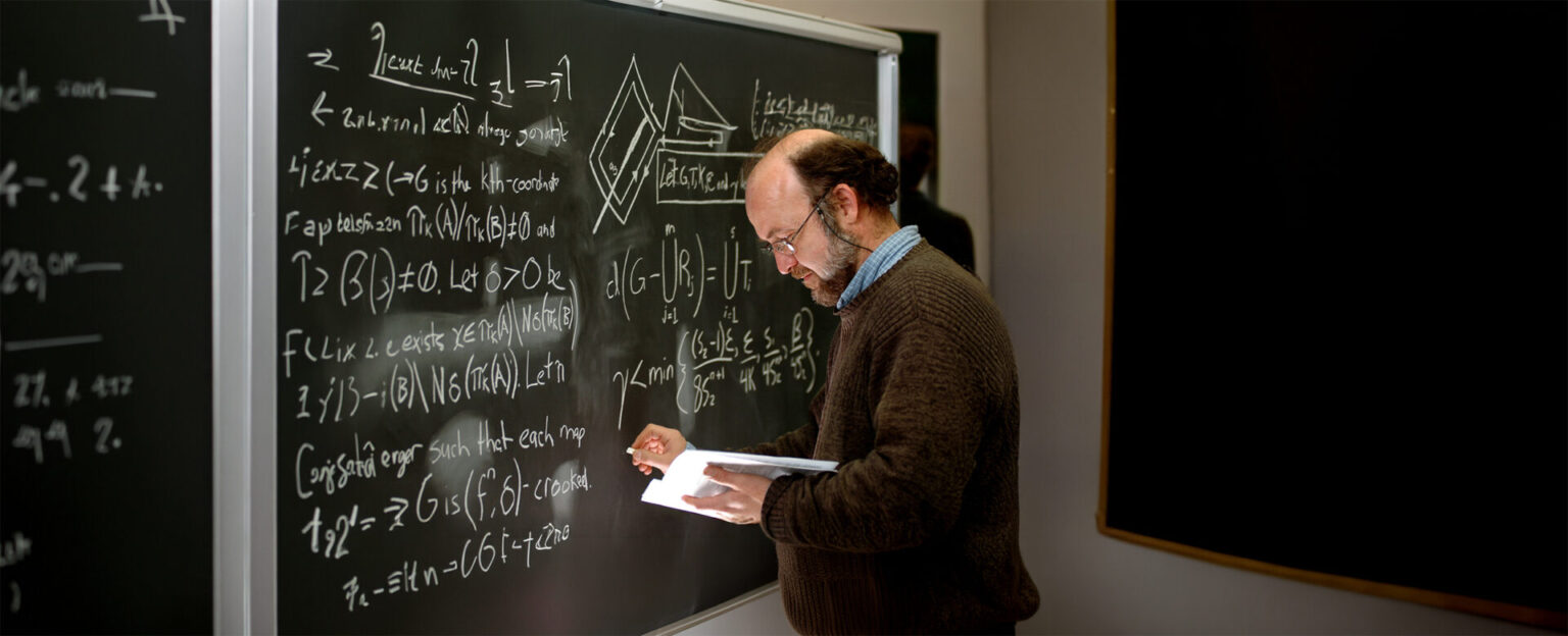 A professor in brown sweater writing on a black board with white equations
