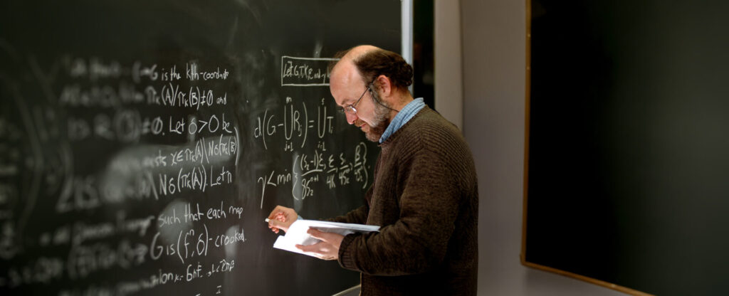 A professor in brown sweater writing on a black board with white equations