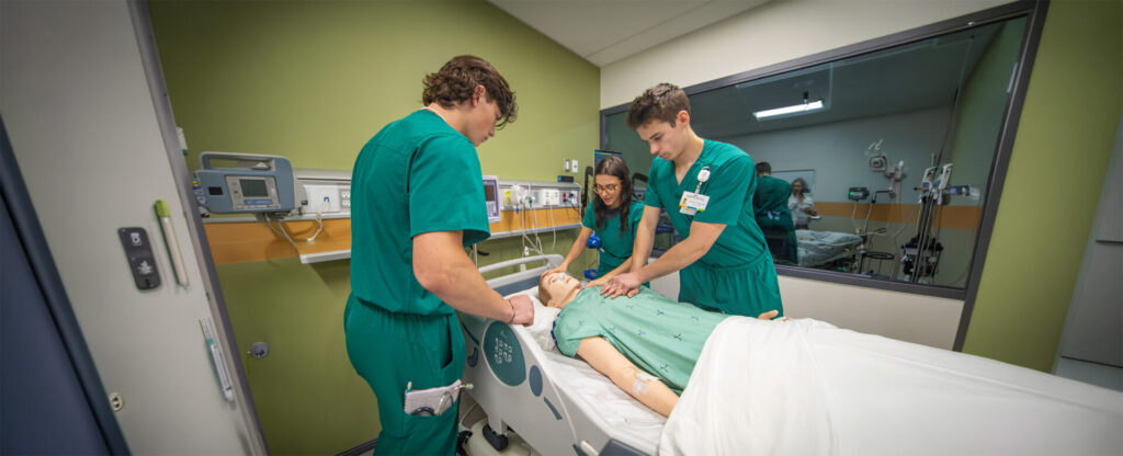 3 nursing students in green scrub all focused on the patient in the simulation lab