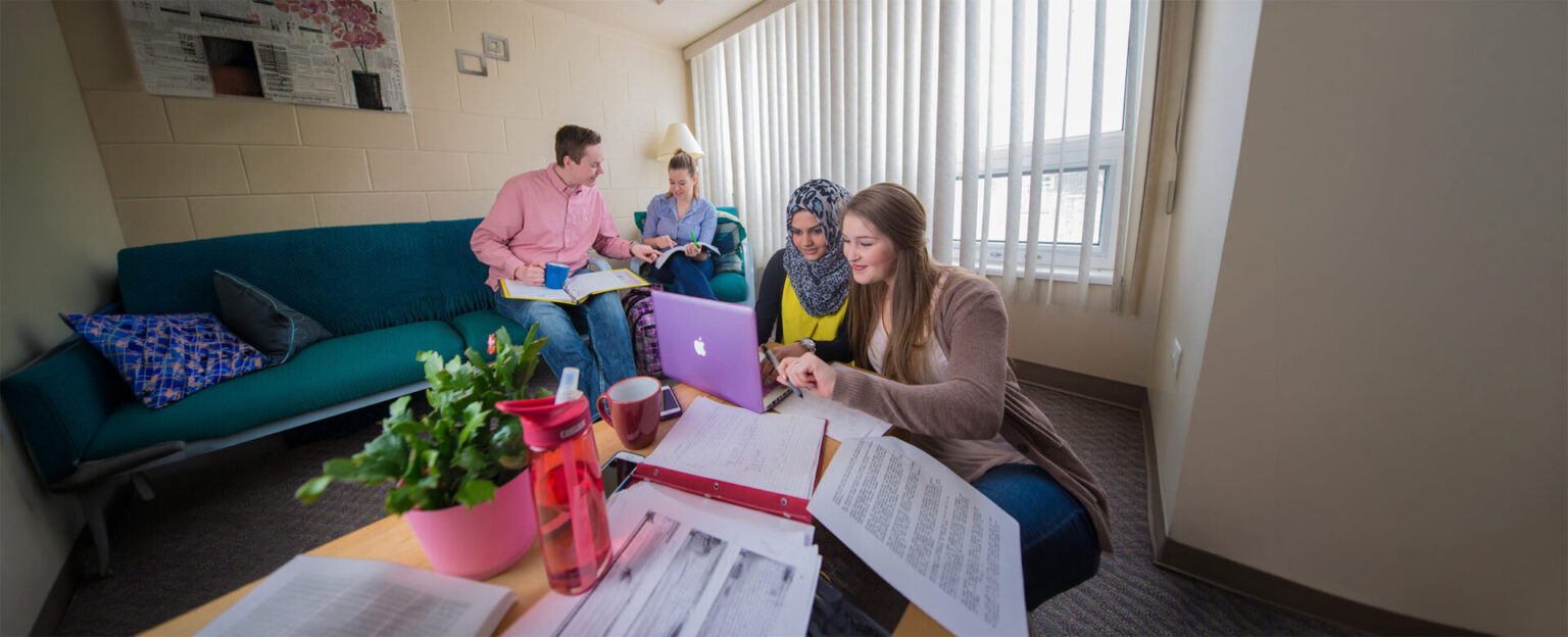 Students studying in the dorm with 2 girls in the forefront studying on a rounded table with their laptop and 2 in the background
