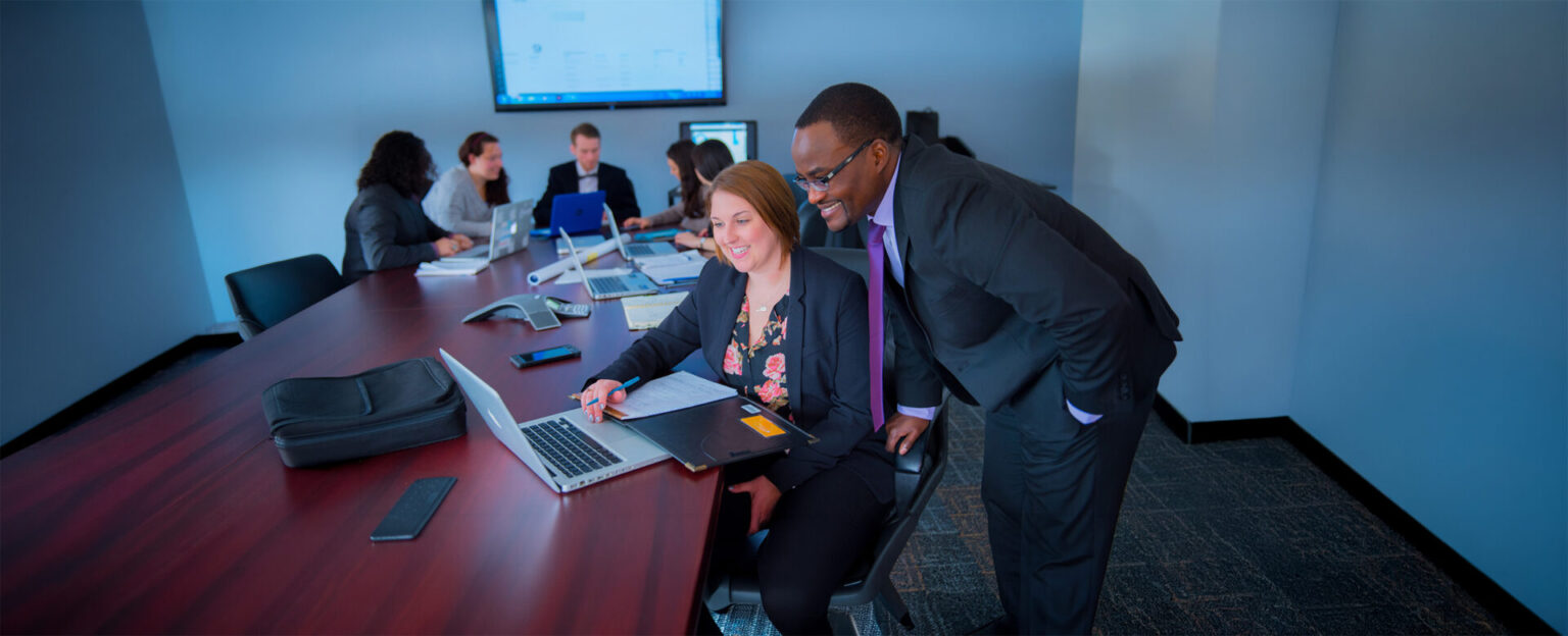 A female student and a professor in business suits looking at a laptop while a group of people are working in the background