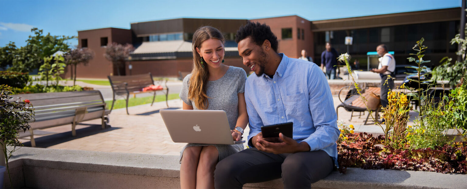2 students working on their laptop and ipad in front of the Nipissing campus entrance