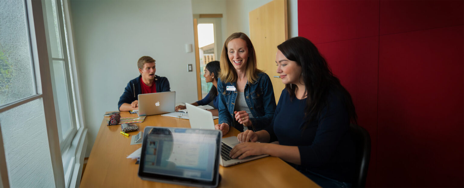2 students working on something on a laptop while 2 other students are in the background behind them
