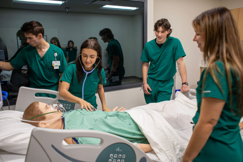 4 nursing students in green scrub, standing around a simulation bed with a robot patient
