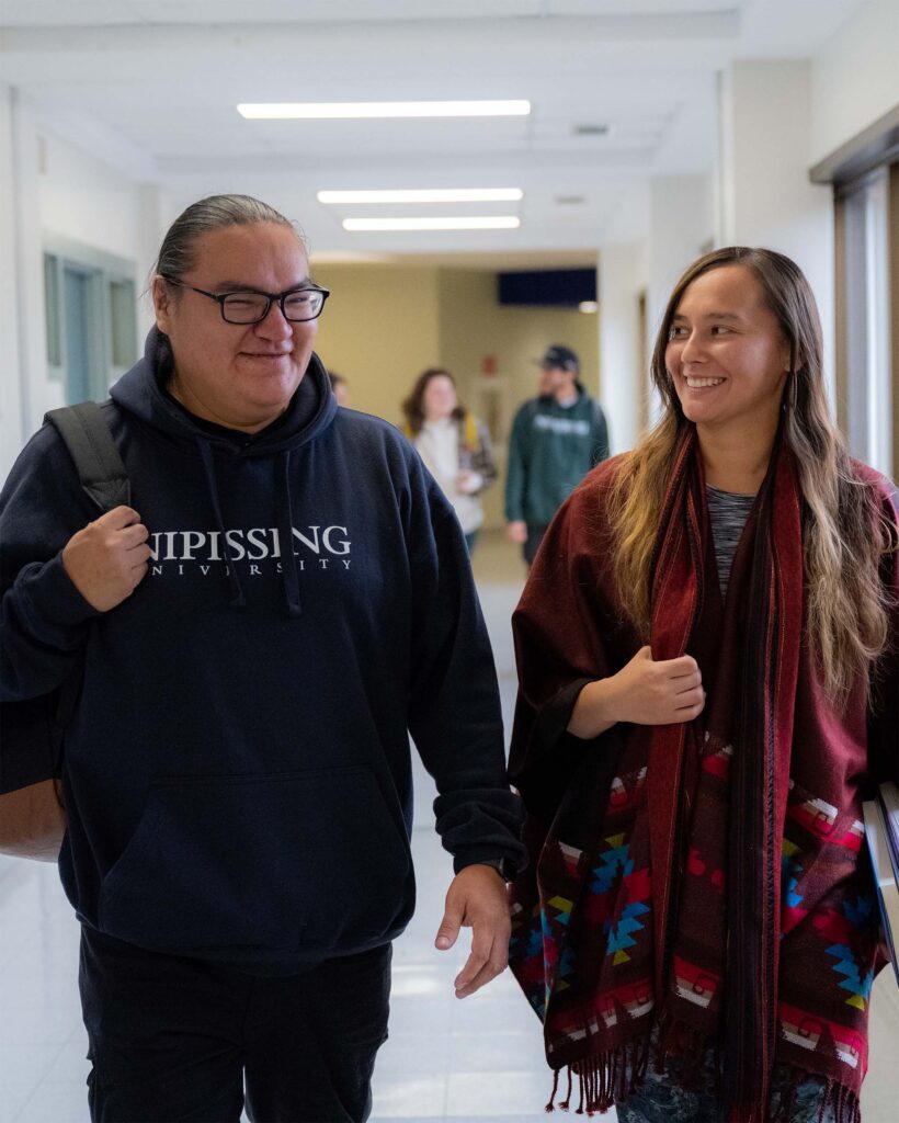 A male student in a navy hoodie and a female student in traditional indigenous clothing walking through the hallway