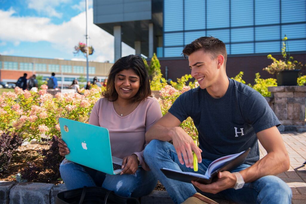 2 students are studying on a laptop with a bush and the library in the background