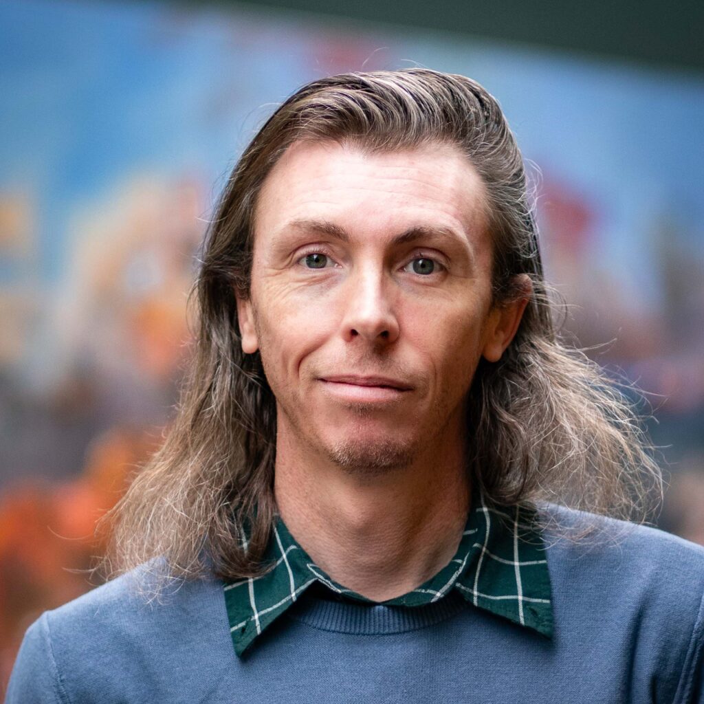 A man with mid-length grey hair against a colorful blurred-out background