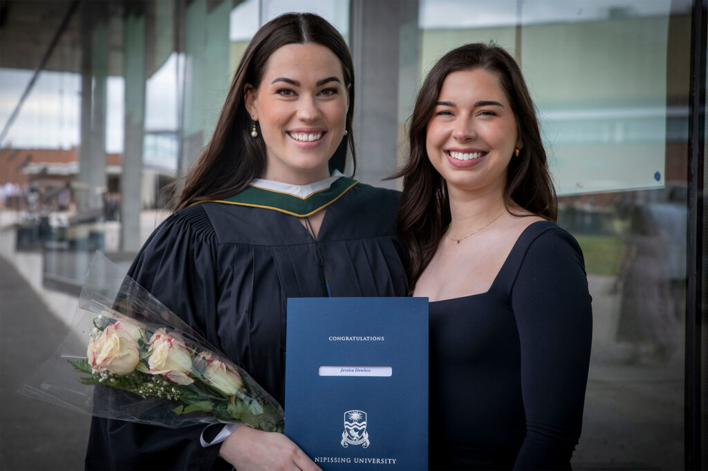 A graduate in gown holding her degrees and flowers next to her friend