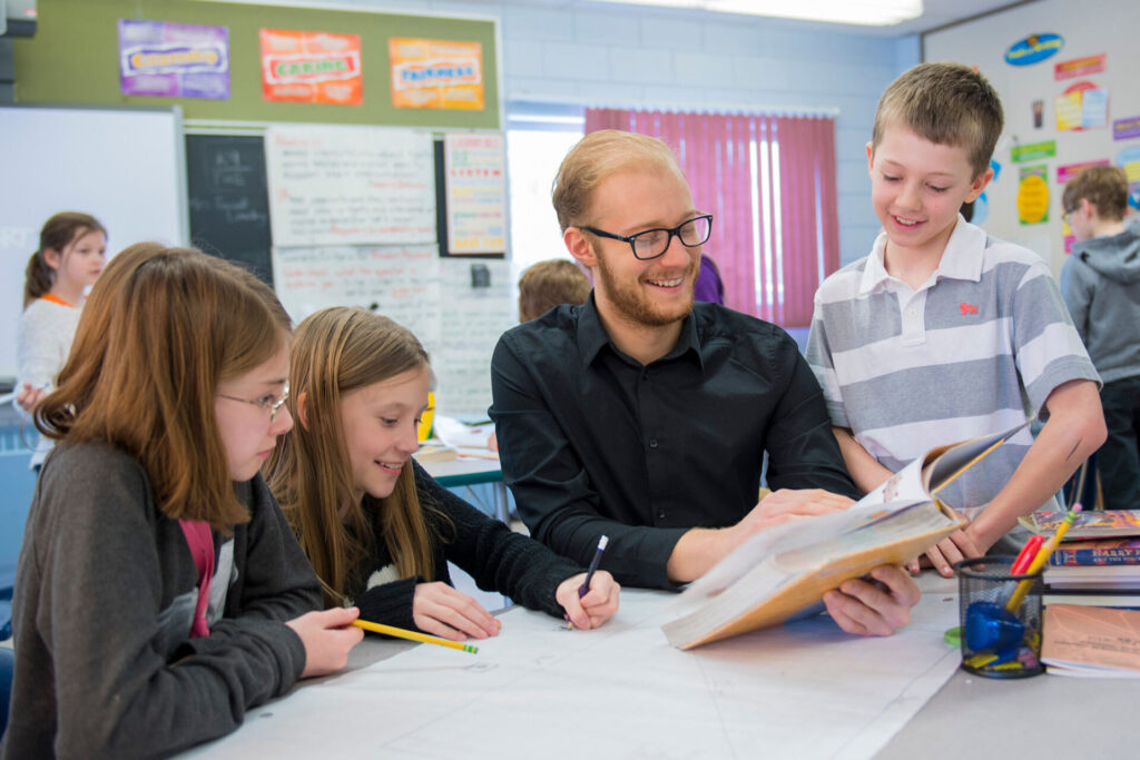 A teacher in black shirt reading a book with a student while 2 other girls sit with them in a class room setting