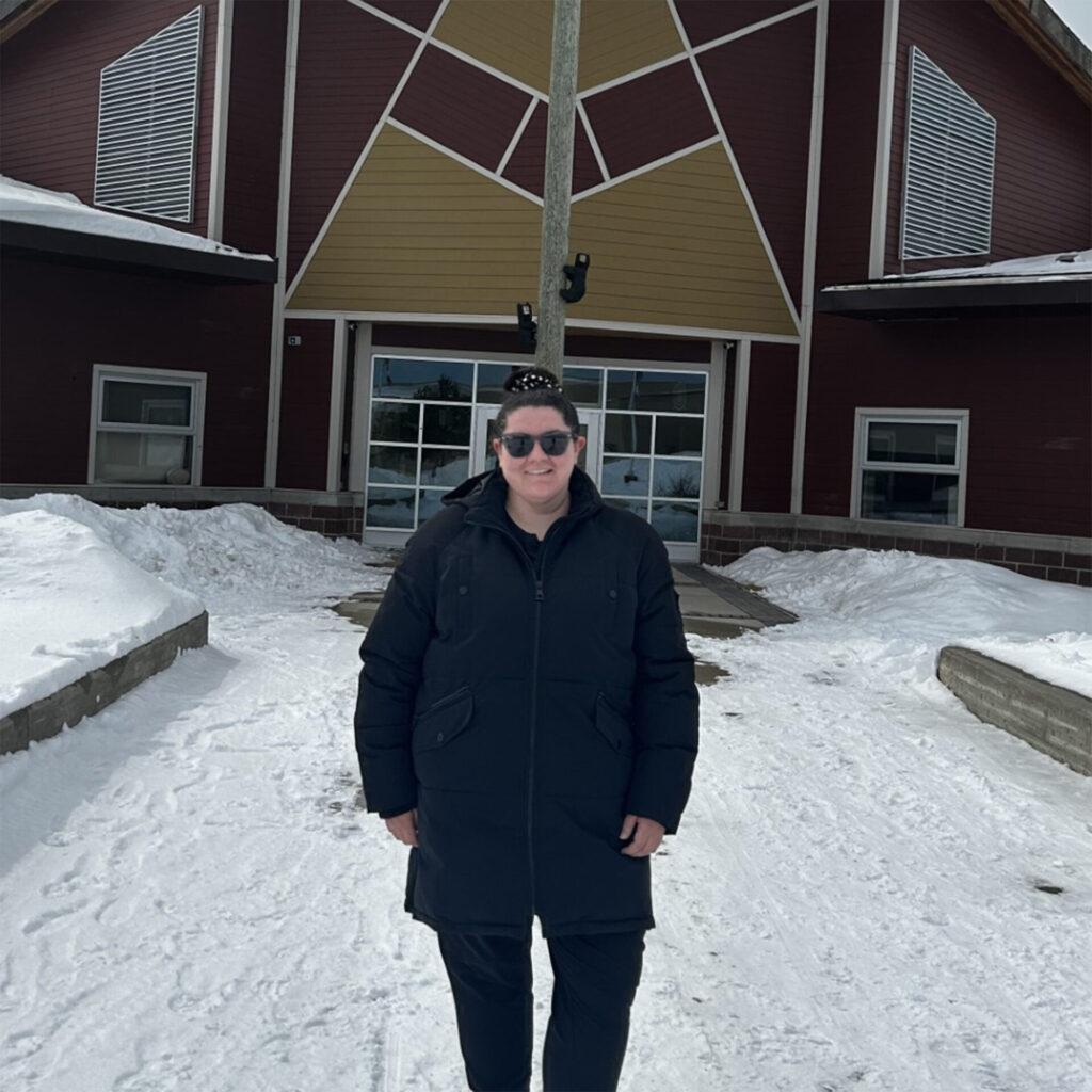 A woman wearing black sunglasses and jacket standing outside in the snow in front of a building