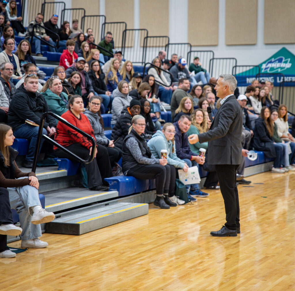Nipissing's president is giving a speech in the gym to a big group of students and parents