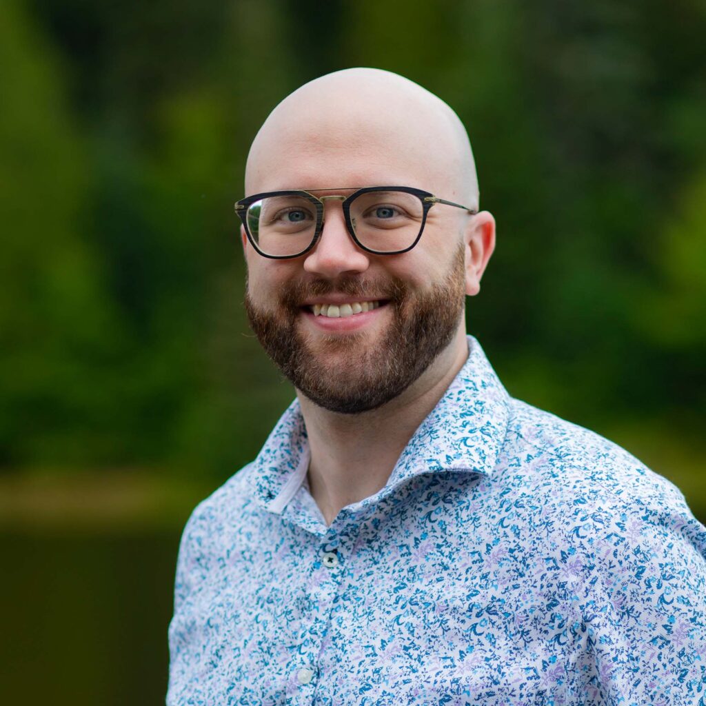 A man in glass wearing a white and blue pattered shirt in front of a blurred nature background