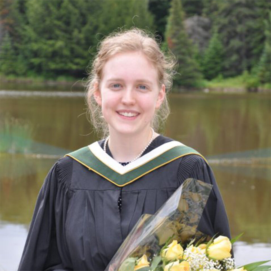 A graduate in convocation gown holding flowers in front of the lake