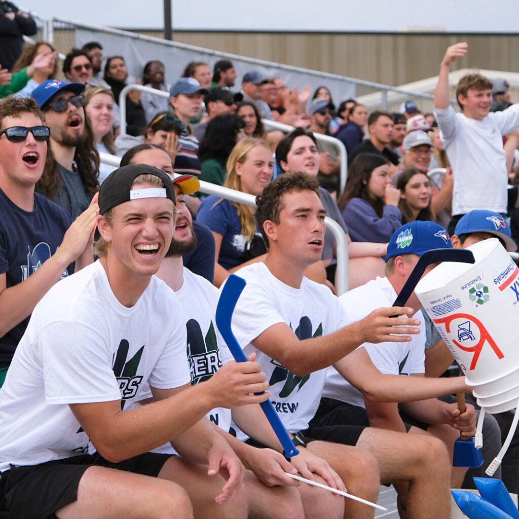 A group of students cheering at a homecoming game on the benches with people in the background