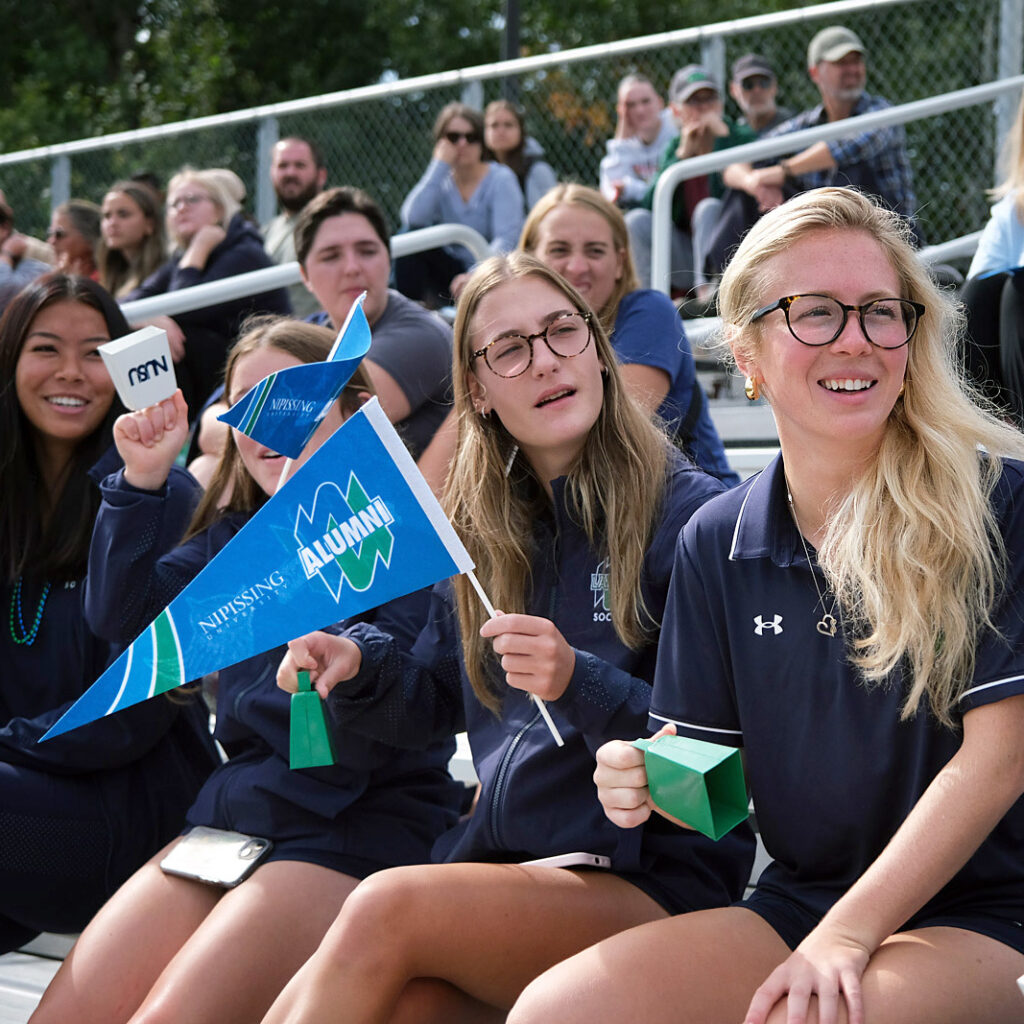 A bench of students holding homecoming flags with people in the background watching a game