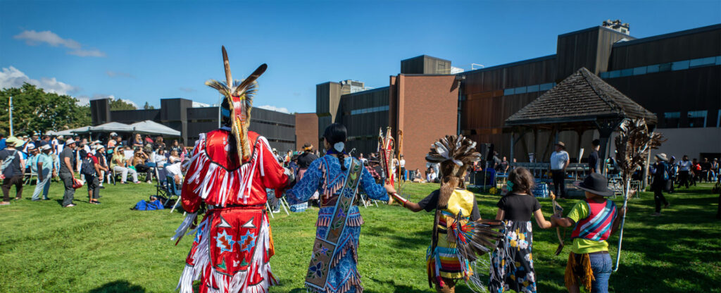 4 people in traditional Indigenous clothing facing the pow wow celebration outdoors