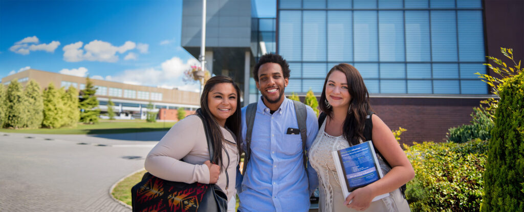 A male student in blue shirt between 2 female students standing outside with the library building behind them in the background