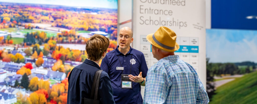 A professor in navy polo talking to a prospective student and his dad at the Ontario University Fair