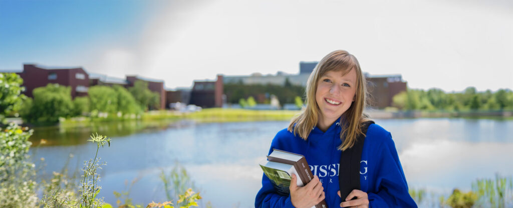 A student in blue Nipissing hoodie holding some books in front of the pond and Nipissing building