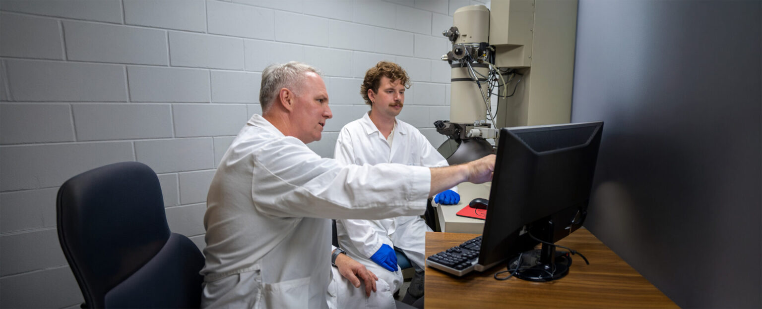 A professor and a student in white lab coat discussing something on a monitor screen in a lab setting